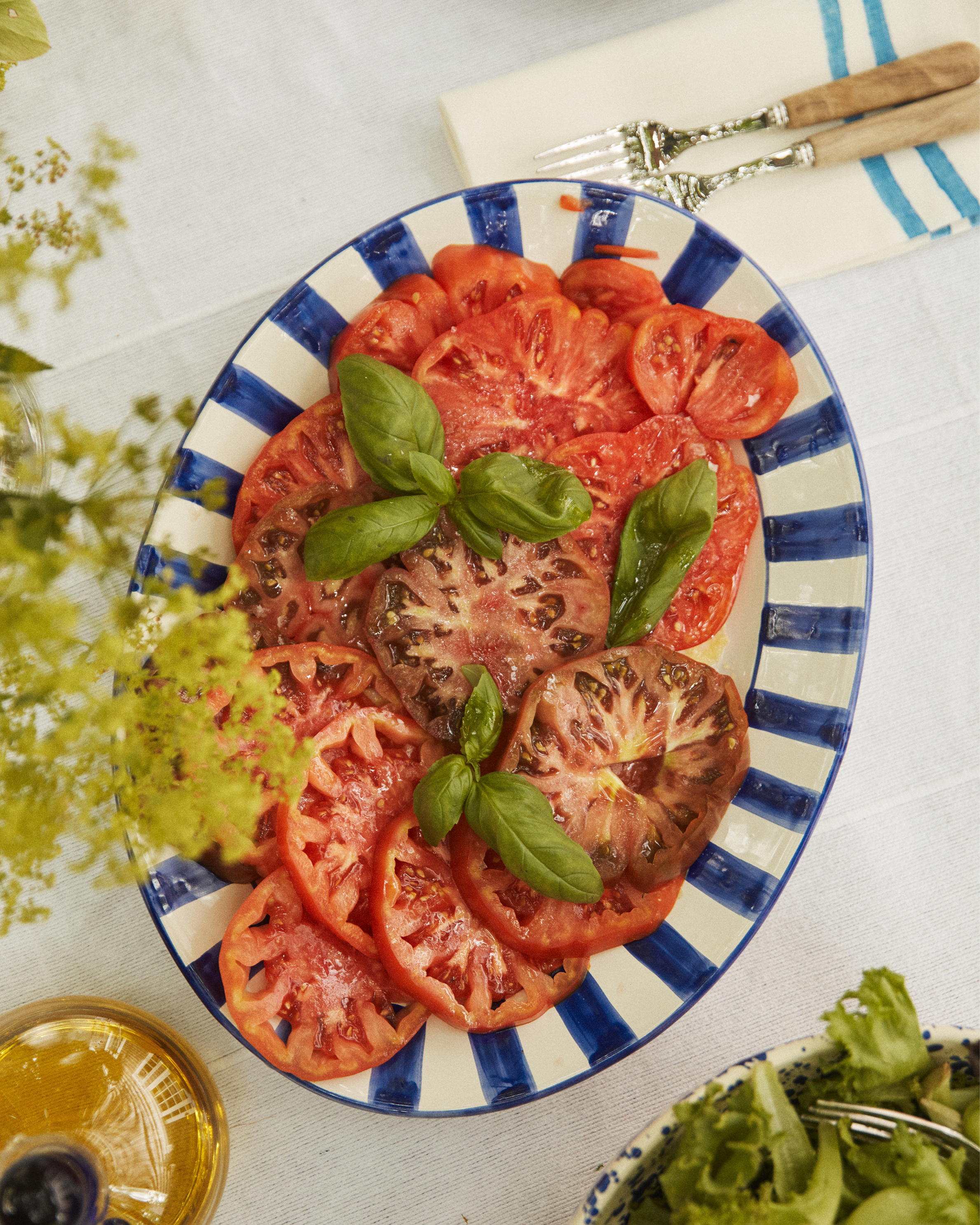 Sliced tomatoes with fresh basil are arranged on the Julius Oval Serving Platter, which features hand-painted blue and white stripes, set on a table alongside cutlery and a salad.