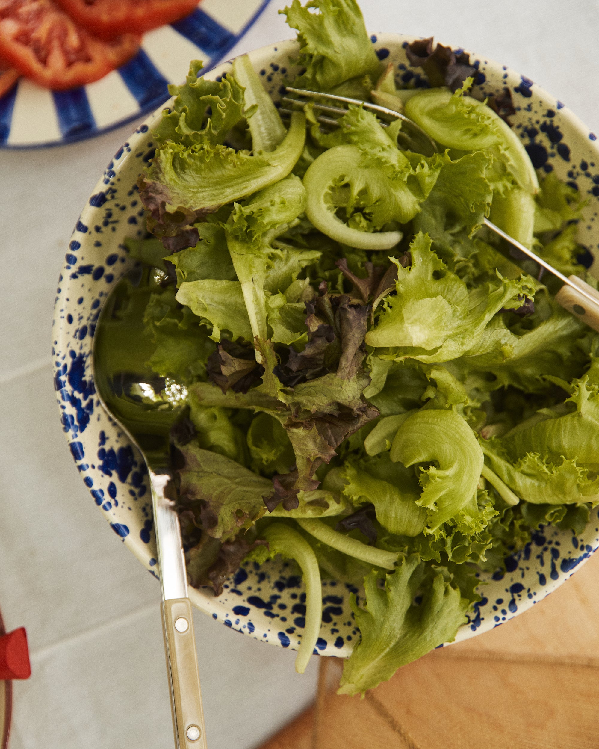 A bowl of mixed green salad is served in the Rita Bowl Splatter, with metal salad servers, beside a plate of sliced tomatoes.