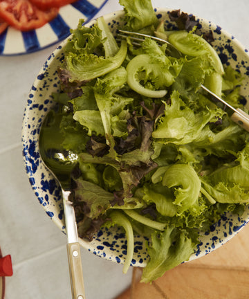 A bowl of mixed green salad is served in the Rita Bowl Splatter, with metal salad servers, beside a plate of sliced tomatoes.
