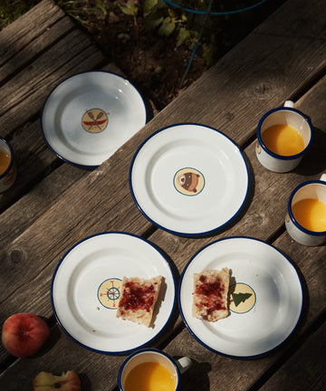 Four camp plates are arranged on a wooden surface in sunlight, along with cups of orange juice, apples, and bread with jam—perfect kids tableware for breakfast.