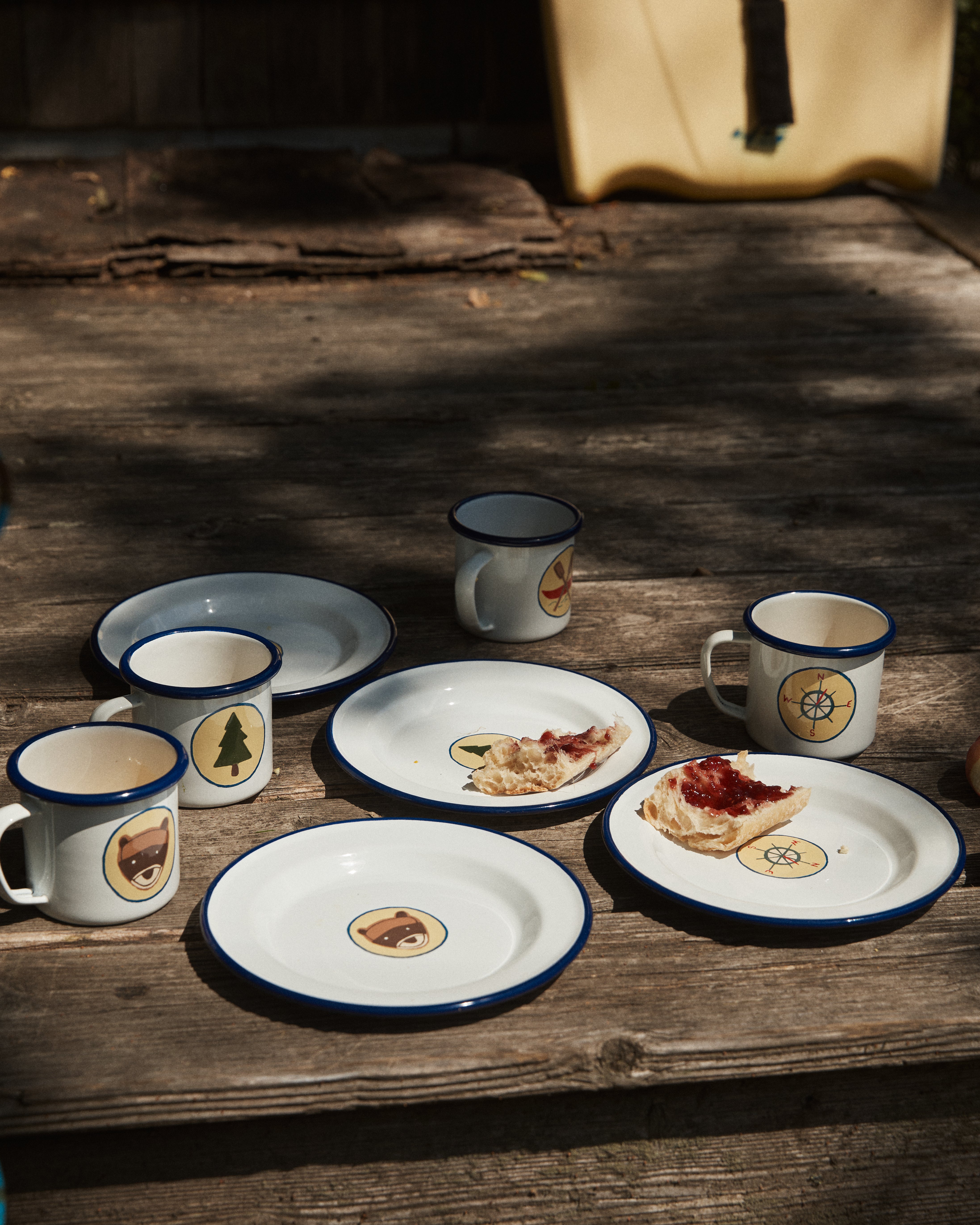 Four Camp Cups, set of 4, with vintage-style bear, tree, and compass designs sit on wooden steps next to enamel plates. Two plates hold bread with red jam, creating a charming kids tableware display.