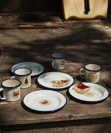 Four Camp Cups, set of 4, with vintage-style bear, tree, and compass designs sit on wooden steps next to enamel plates. Two plates hold bread with red jam, creating a charming kids tableware display.