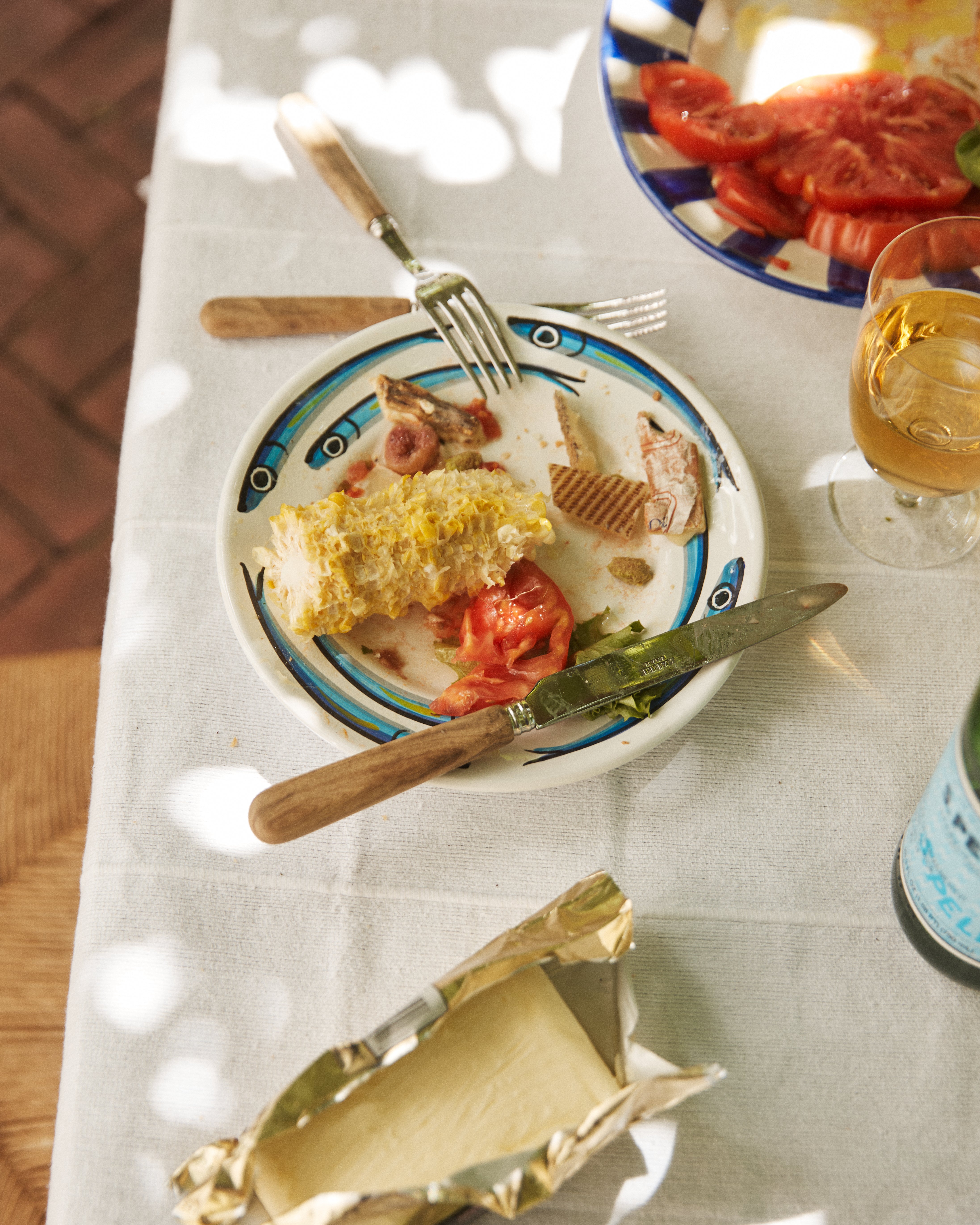 A partially eaten corn cob, tomato slices, and bits of meat rest on a Tommaso Small Plate. Utensils, a drink, extra tomatoes, and an open butter dish inspired by the Amalfi Coast complete the scene. Set includes 4 plates.