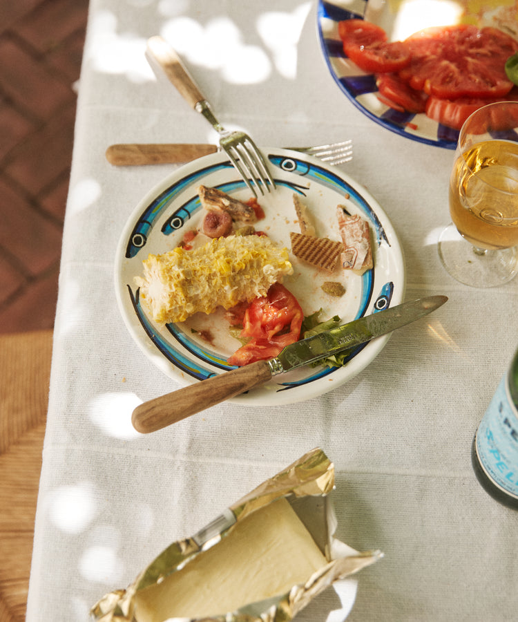 A partially eaten corn cob, tomato slices, and bits of meat rest on a Tommaso Small Plate. Utensils, a drink, extra tomatoes, and an open butter dish inspired by the Amalfi Coast complete the scene. Set includes 4 plates.