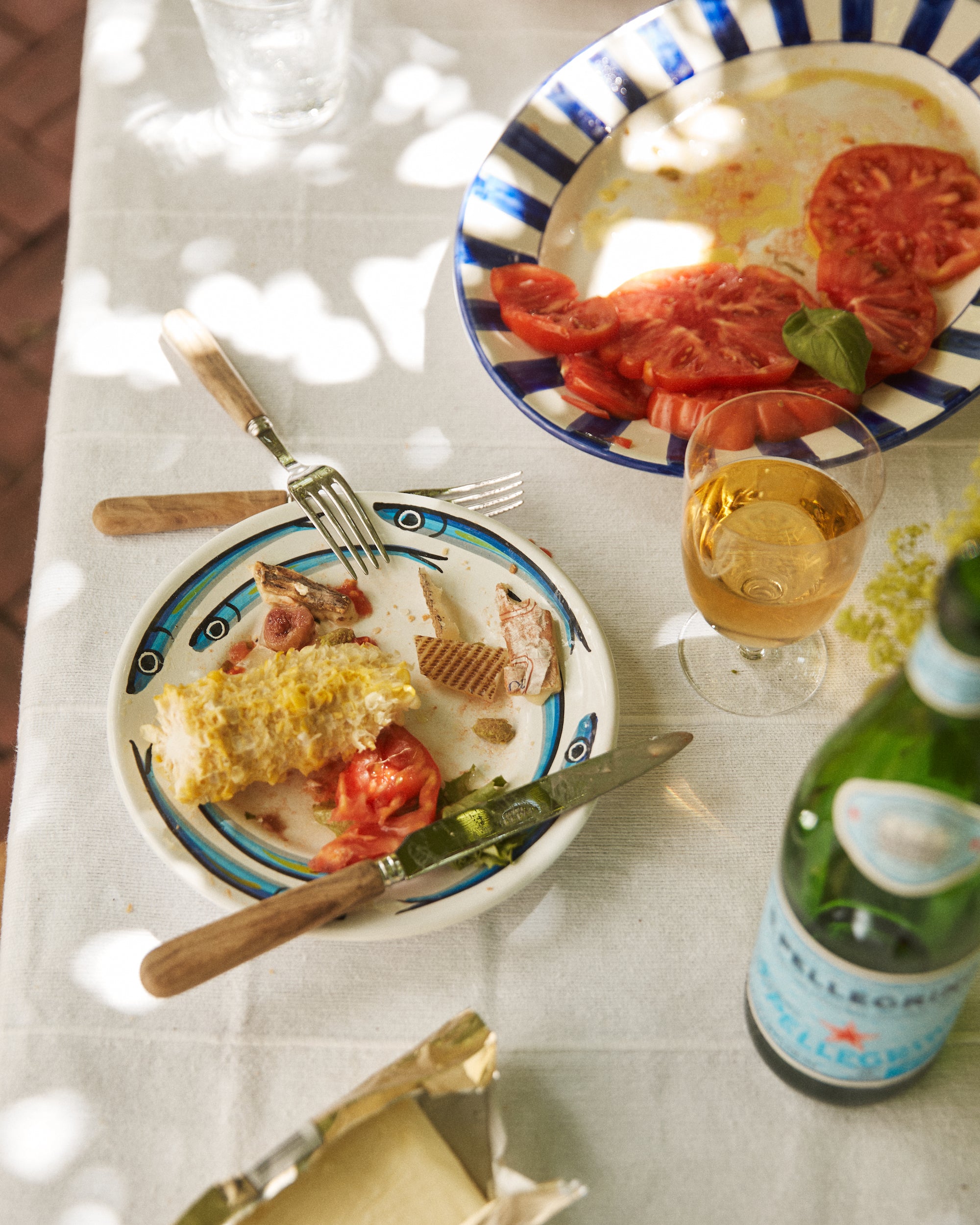 A partially eaten meal on a white tablecloth, with corn on the cob, fish, tomatoes, and wine served on vibrant Tommaso Small Plates (Set of 4), featuring hand-painted designs inspired by the Amalfi Coast.