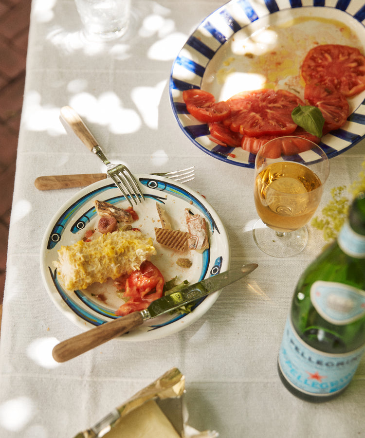 A partially eaten meal on a white tablecloth, with corn on the cob, fish, tomatoes, and wine served on vibrant Tommaso Small Plates (Set of 4), featuring hand-painted designs inspired by the Amalfi Coast.
