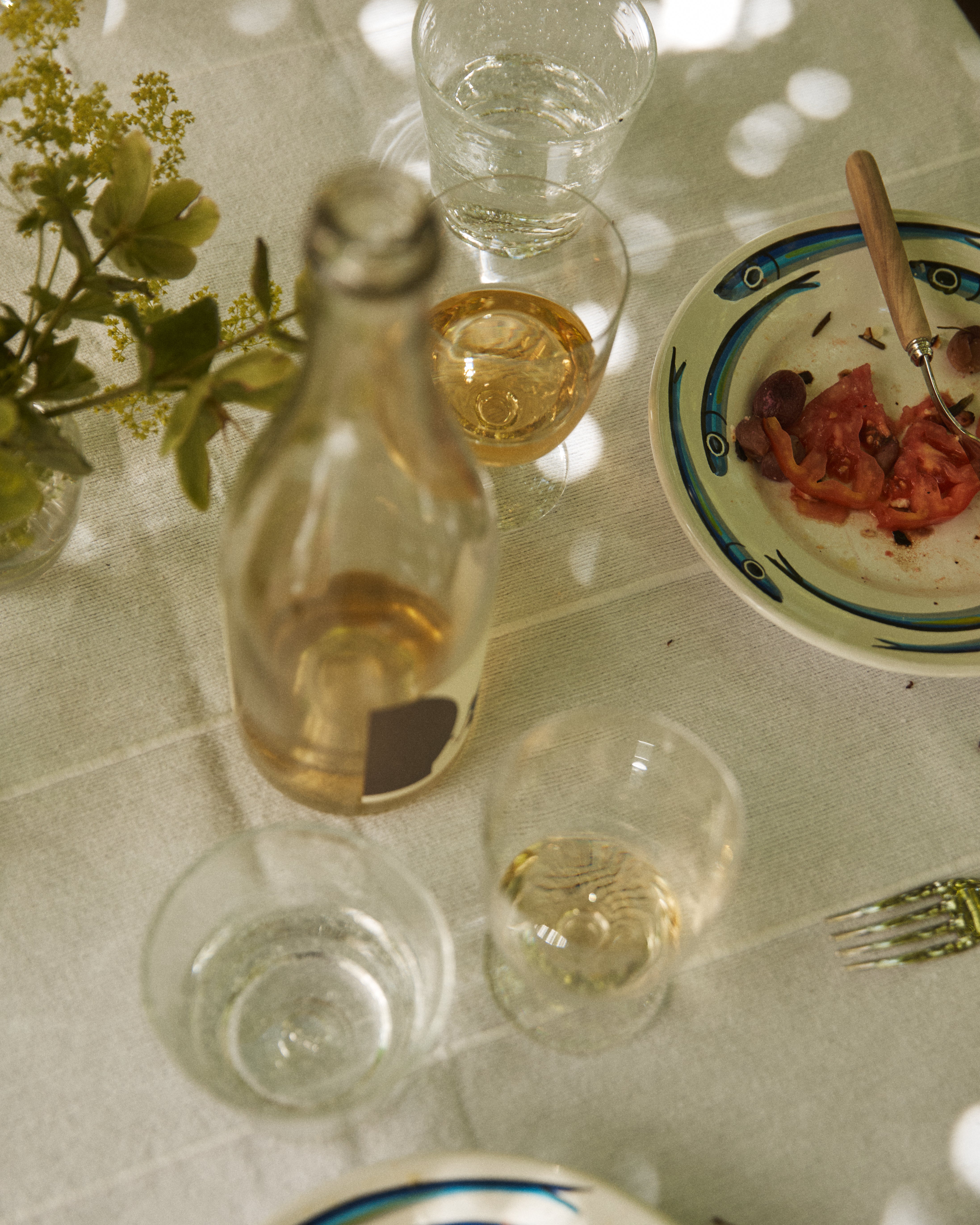 A table set with French glassware—including a Lauren Water Glass—a clear bottle, wine glass, plate with tomato and olives, empty fork, and a small vase of flowers on a white tablecloth.