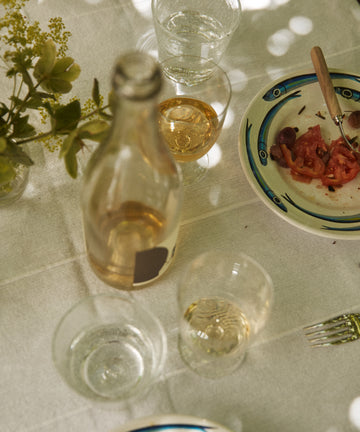A table set with French glassware—including a Lauren Water Glass—a clear bottle, wine glass, plate with tomato and olives, empty fork, and a small vase of flowers on a white tablecloth.