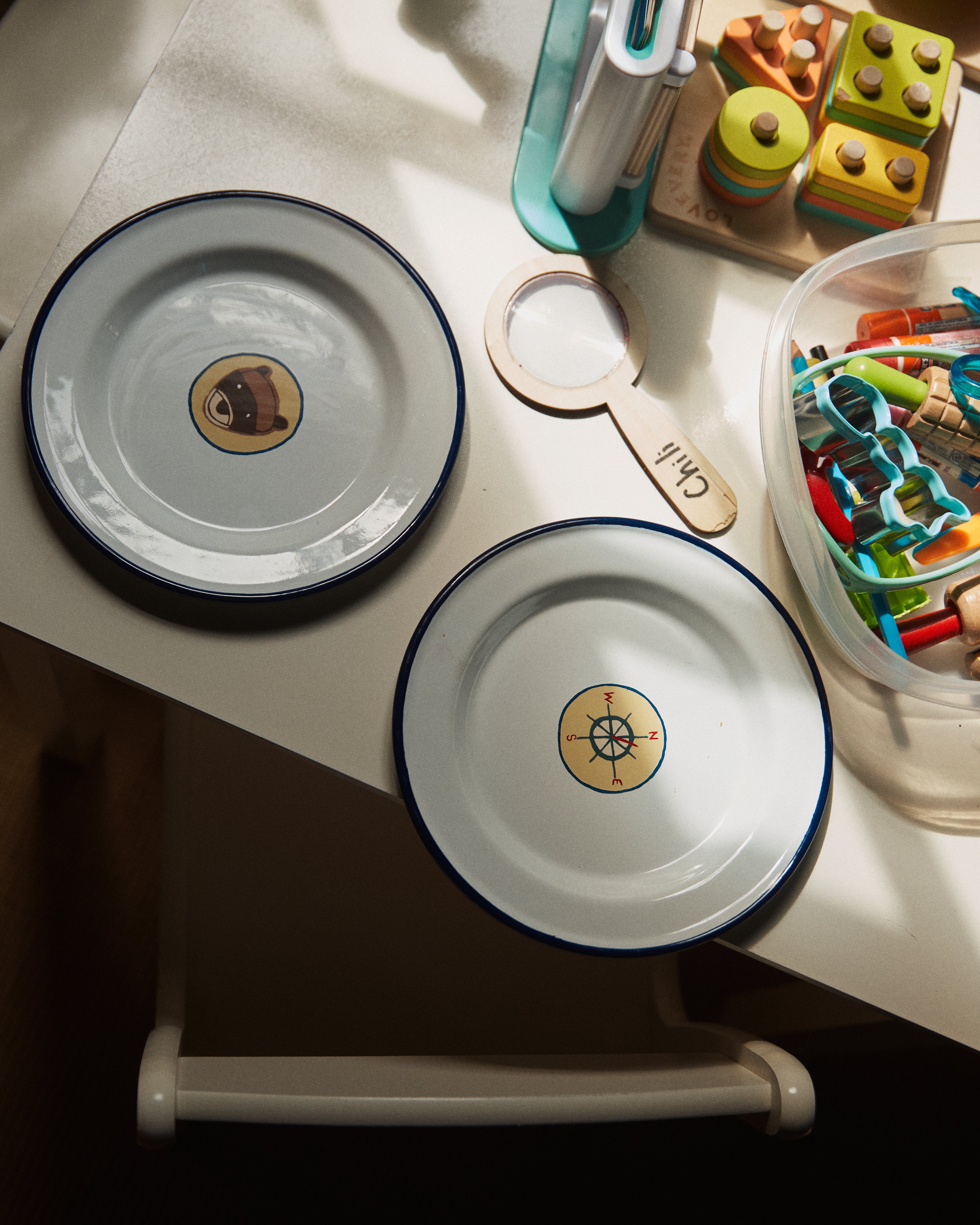 A set of four lightweight, durable Camp Plates with blue rims—featuring avocado and compass designs—are displayed on a table amid colorful toys, craft materials, and vintage scout badges.