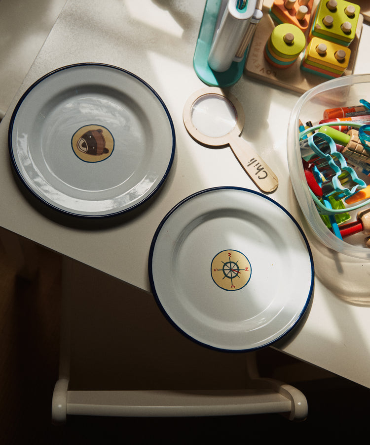 A set of four lightweight, durable Camp Plates with blue rims—featuring avocado and compass designs—are displayed on a table amid colorful toys, craft materials, and vintage scout badges.