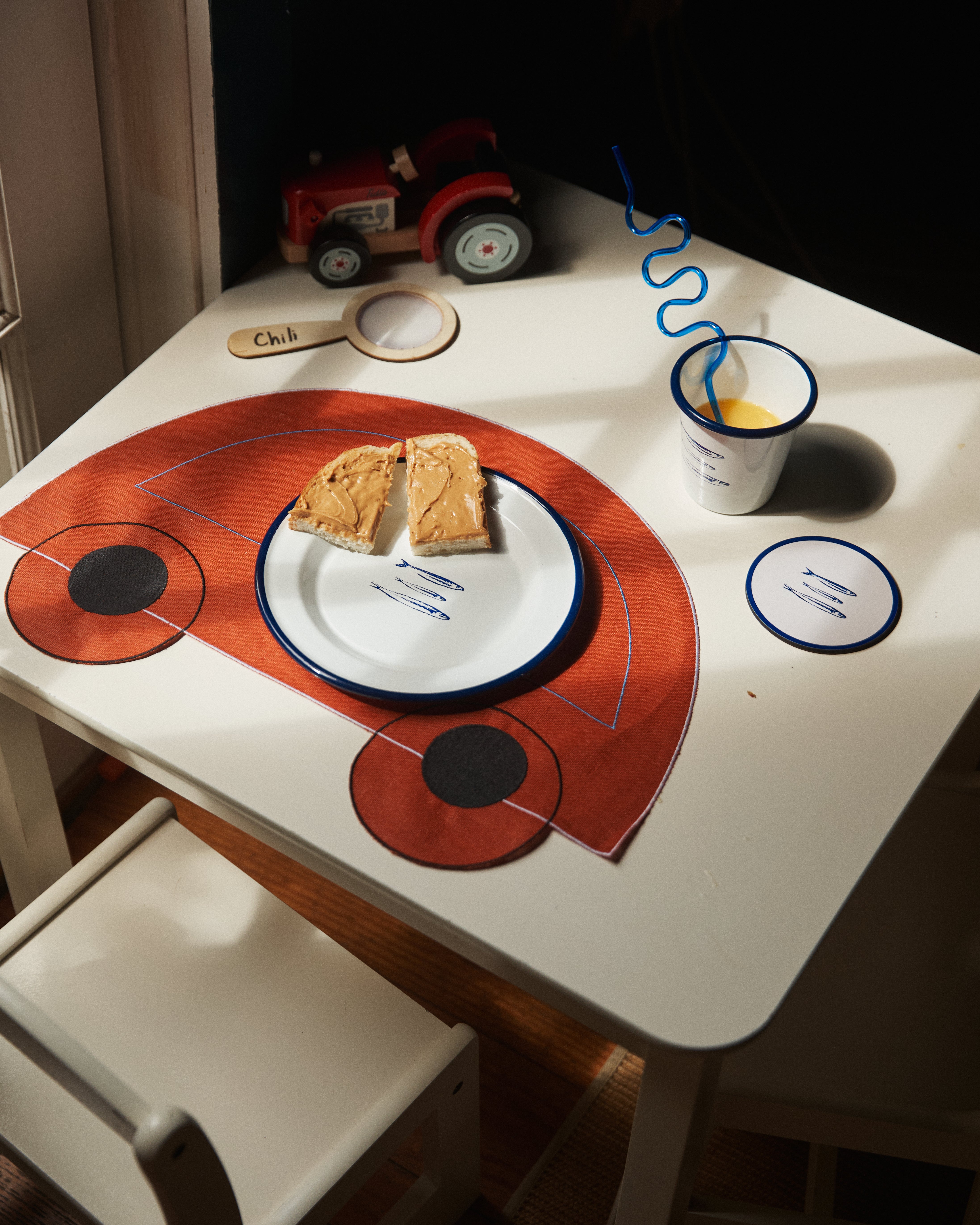 A small table with a plate of peanut butter toast, orange juice with a curly straw, a toy tractor, and a Thomas Placemat (Set of 4) for kids.