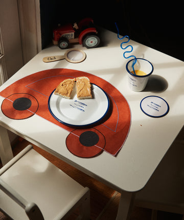 A small table with a plate of peanut butter toast, orange juice with a curly straw, a toy tractor, and a Thomas Placemat (Set of 4) for kids.