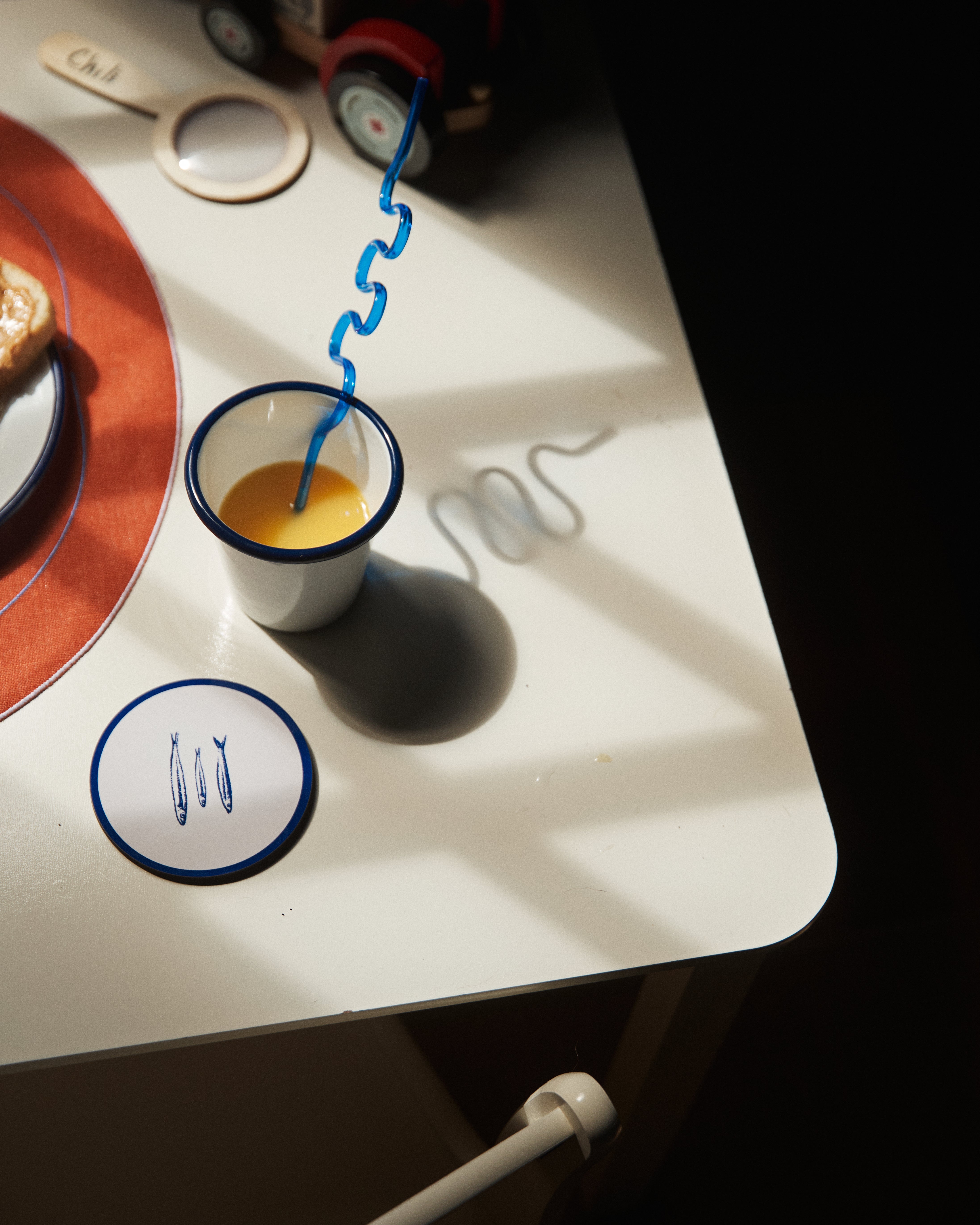 A cup of orange juice with a curly blue straw sits on a white table in sunlight beside Costa Coaster, Set of 6, a saucer, toast, a toy train, and utensils.