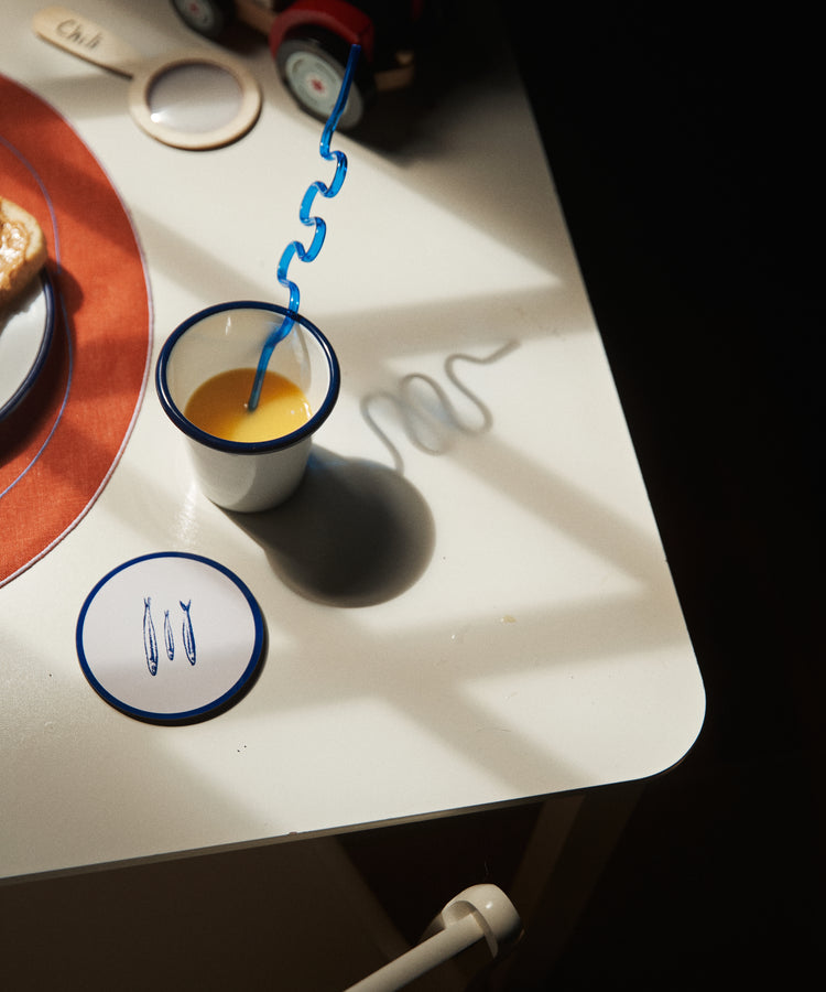 A cup of orange juice with a curly blue straw sits on a white table in sunlight beside Costa Coaster, Set of 6, a saucer, toast, a toy train, and utensils.
