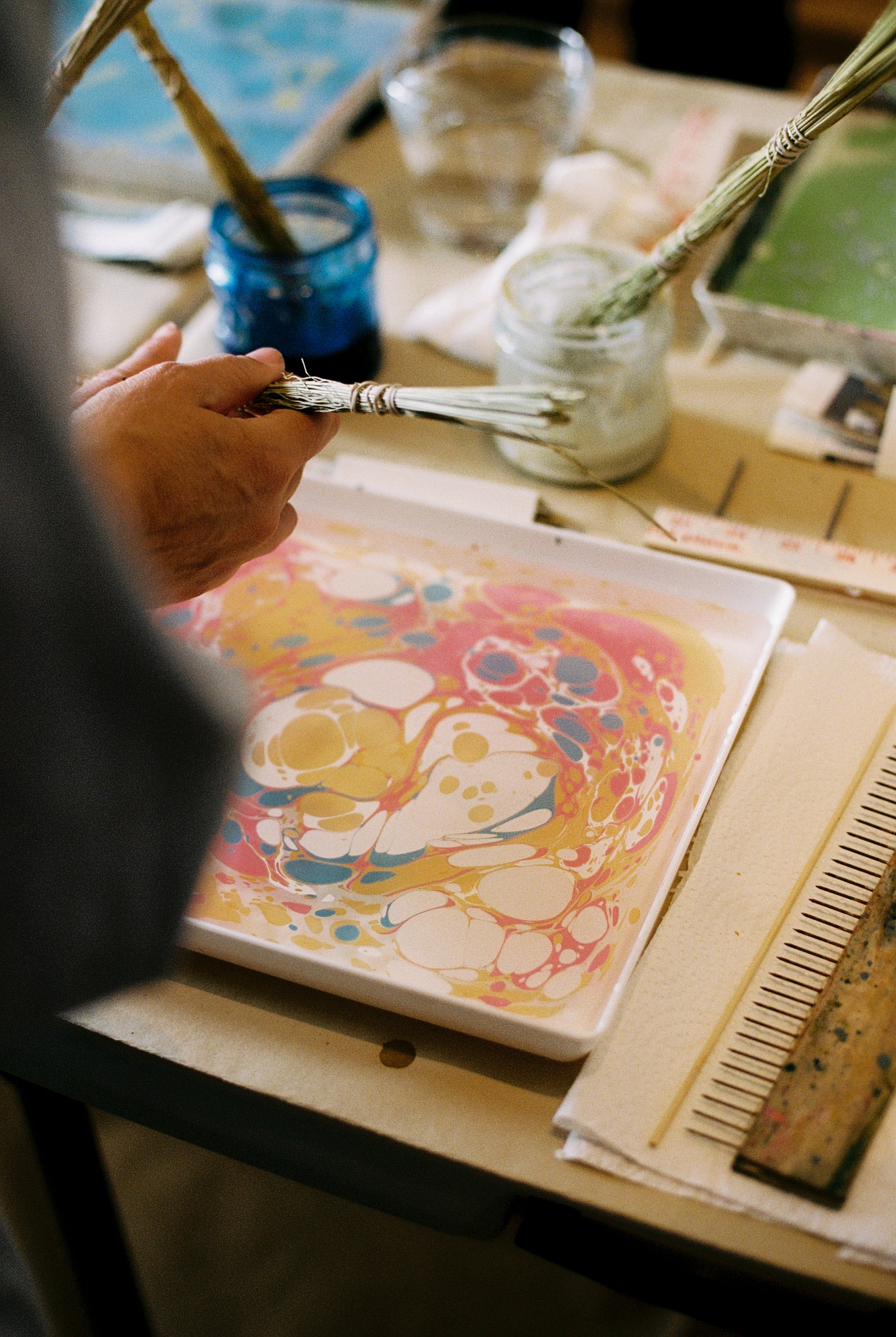 During the PORTA x Ansuini & Co. Marbling Workshop, a participant uses a stick to marble vibrant paint floating on water in a tray—a technique inspired by Florentine artistry—with various art supplies and tools from the workshop arranged on the table nearby.