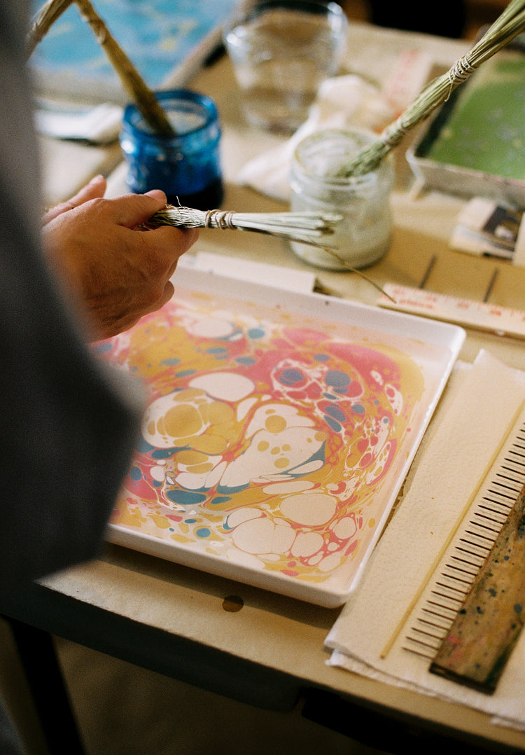 During the Wednesday 30th September, 2026 marbling workshop inspired by Florentine art, a person uses a brush to create colorful patterns on water, with jars, cocktail napkins, and tools arranged on the table.