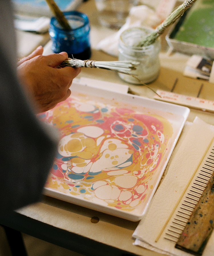 During the PORTA x Ansuini & Co. Marbling Workshop, a participant uses a stick to marble vibrant paint floating on water in a tray—a technique inspired by Florentine artistry—with various art supplies and tools from the workshop arranged on the table nearby.