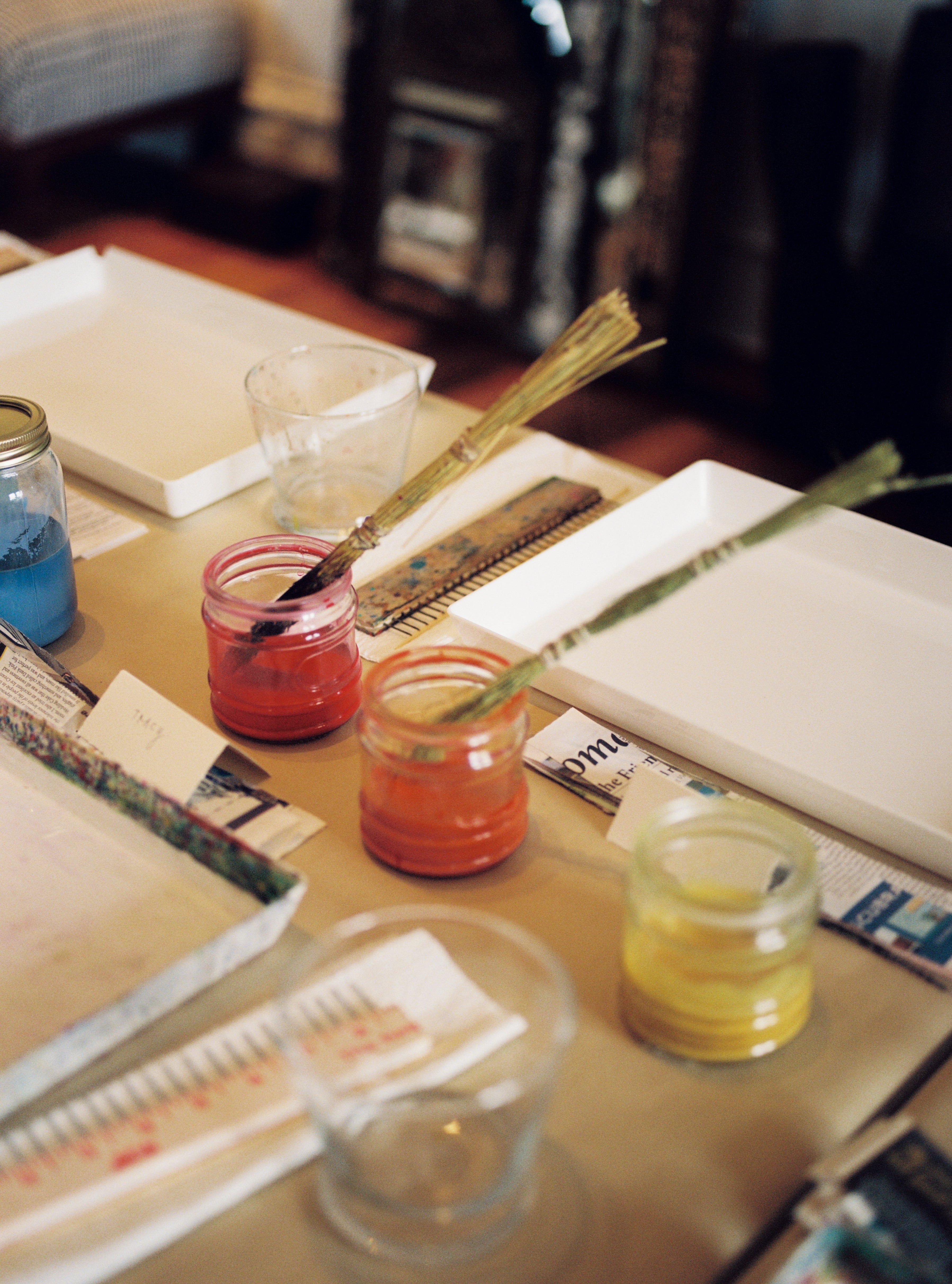 Three jars of paint—red, orange, and yellow—sit on a table with brushes inside. Nearby are trays, rulers, glass cups, and scattered papers from the PORTA x Ansuini & Co. Marbling Workshop inspired by traditional Florentine art.