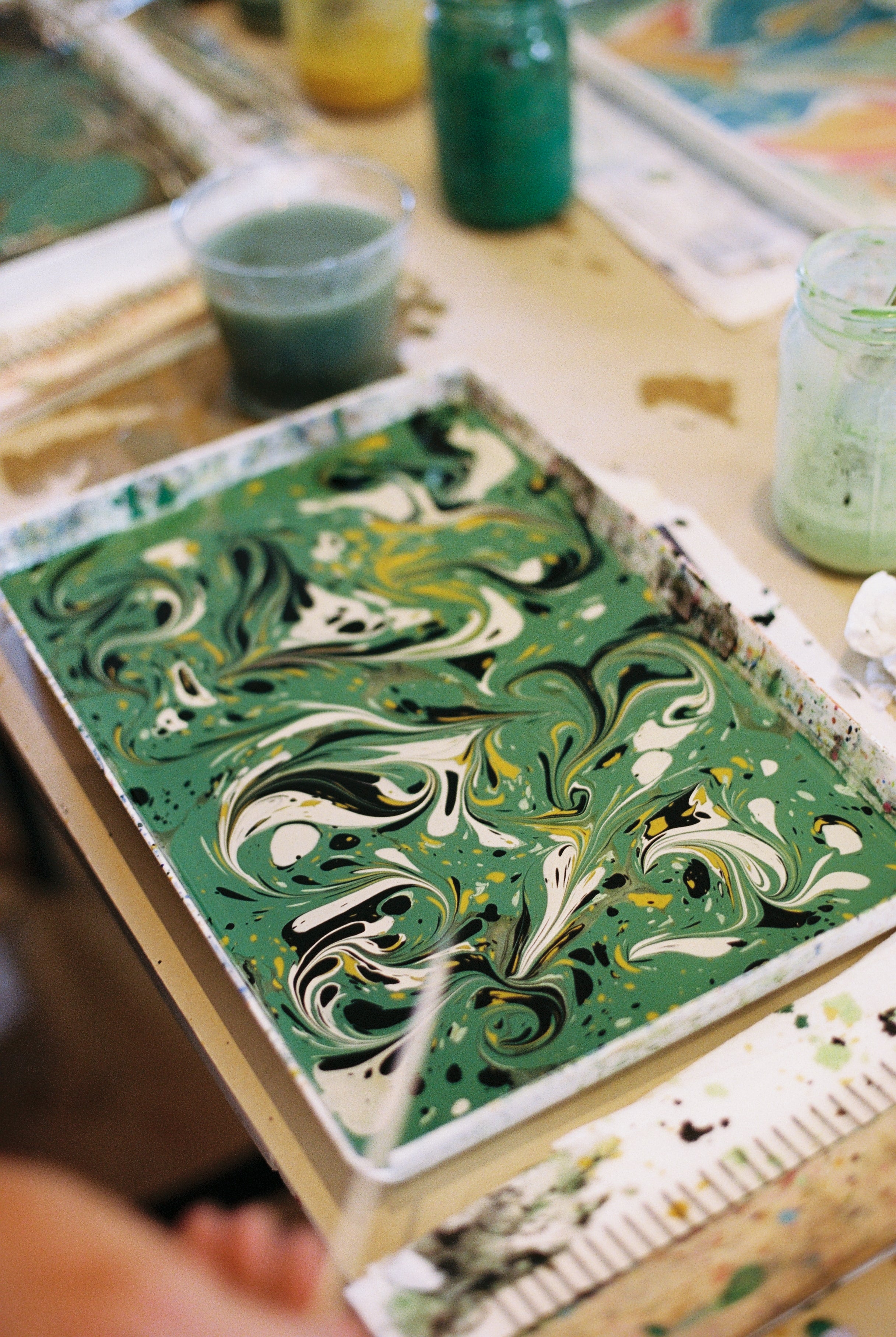 A rectangular tray filled with green, white, yellow, and black marbled paint sits on a table surrounded by jars and art supplies, capturing the essence of Florentine creativity in the PORTA x Ansuini & Co. Marbling Workshop.