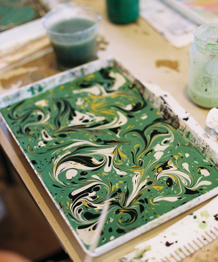 A rectangular tray filled with green, white, yellow, and black marbled paint sits on a table surrounded by jars and art supplies, capturing the essence of Florentine creativity in the PORTA x Ansuini & Co. Marbling Workshop.