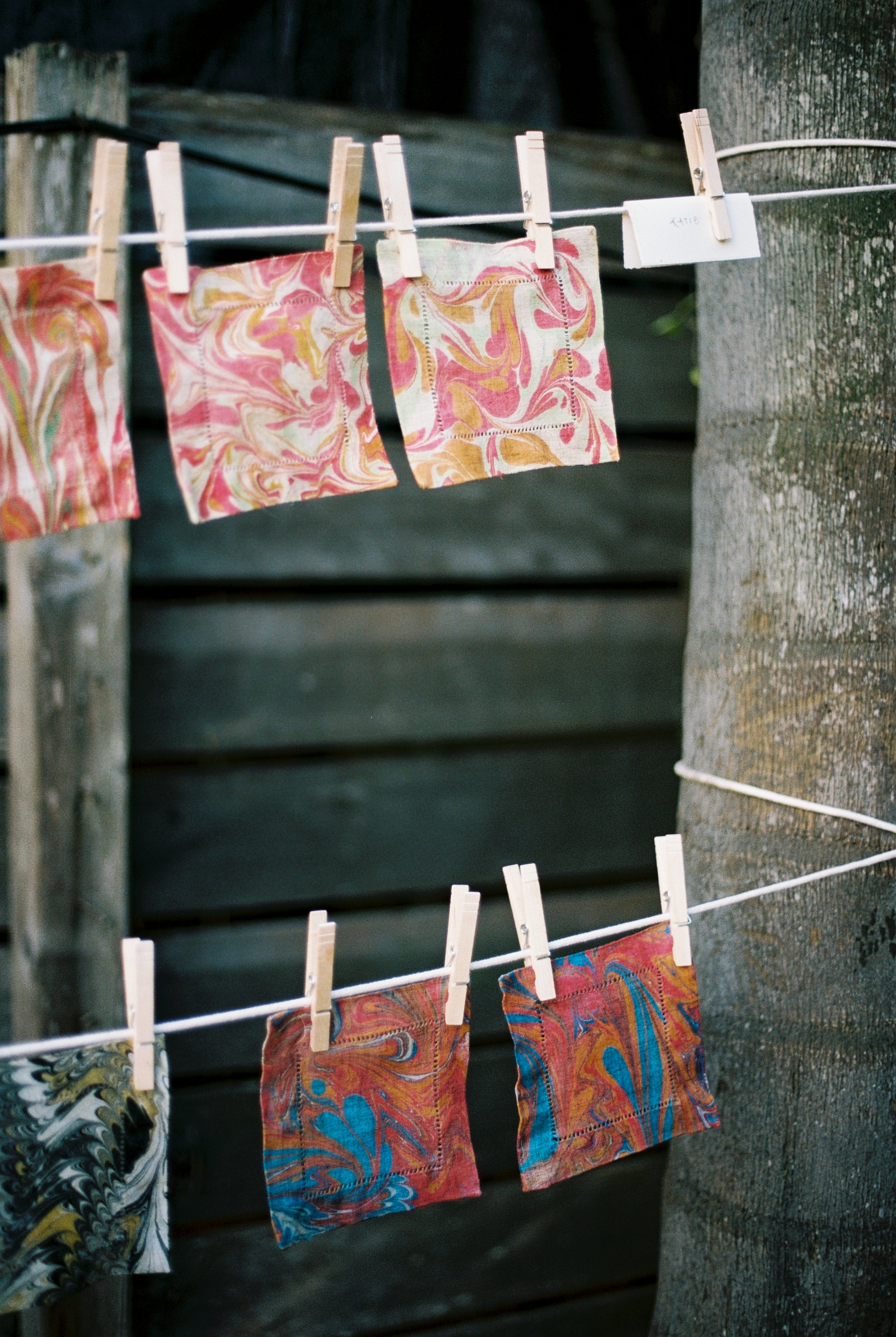 Vivid marbled fabric squares created in the PORTA x Ansuini & Co. Marbling Workshop are strung on clotheslines with wooden clothespins outdoors, displayed against a wooden fence and beside a tree trunk.