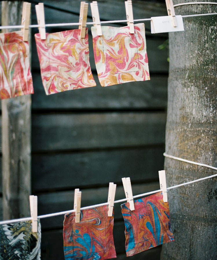 Vivid marbled fabric squares created in the PORTA x Ansuini & Co. Marbling Workshop are strung on clotheslines with wooden clothespins outdoors, displayed against a wooden fence and beside a tree trunk.