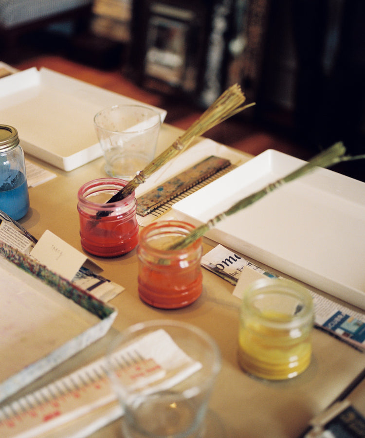 Three jars of paint—red, orange, and yellow—sit on a table with brushes inside. Nearby are trays, rulers, glass cups, and scattered papers from the PORTA x Ansuini & Co. Marbling Workshop inspired by traditional Florentine art.