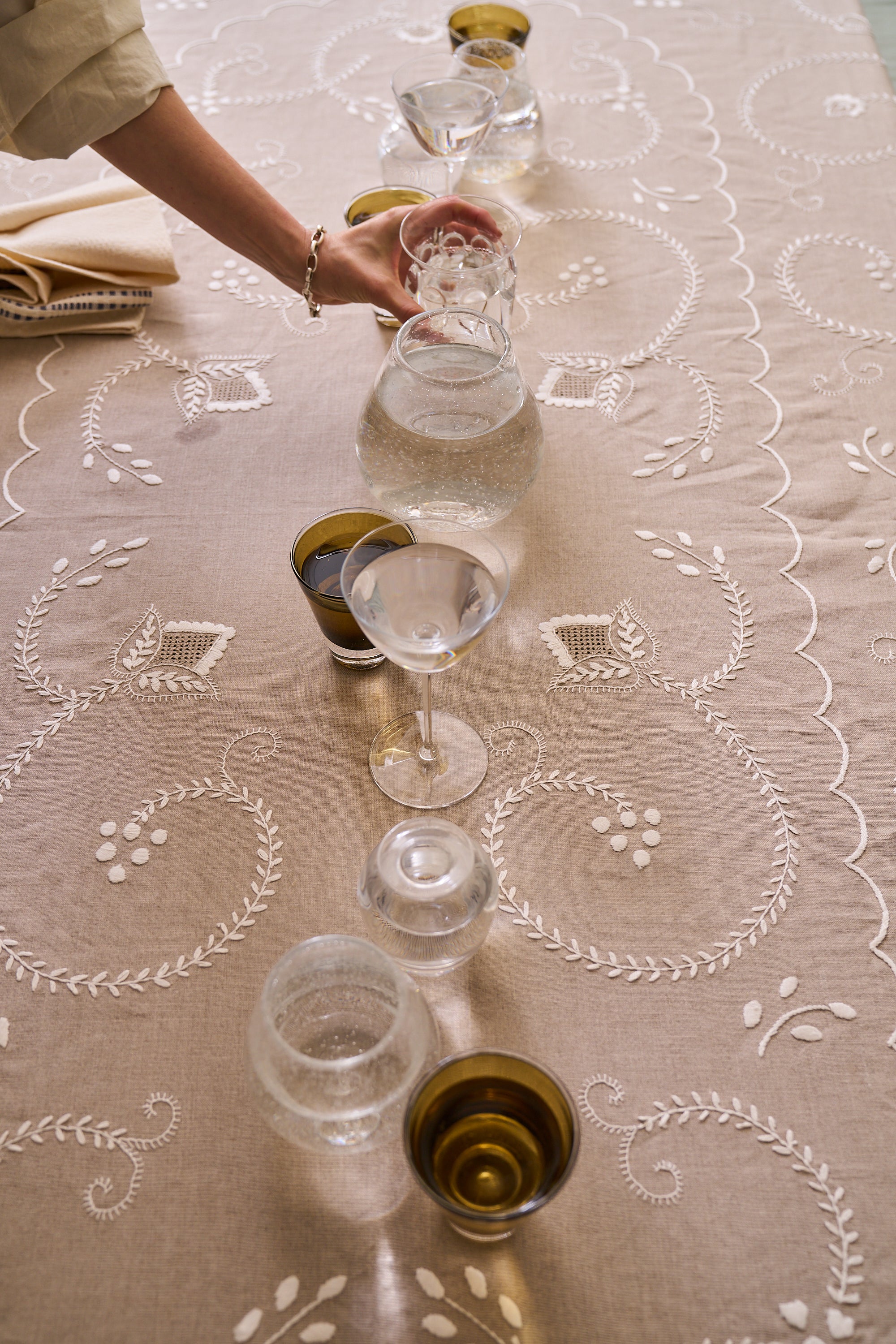 A hand arranges glassware and small cups filled with water on the Inez Tablecloth, crafted from raw linen and hand embroidered with traditional Portuguese designs, with a folded napkin in the corner.