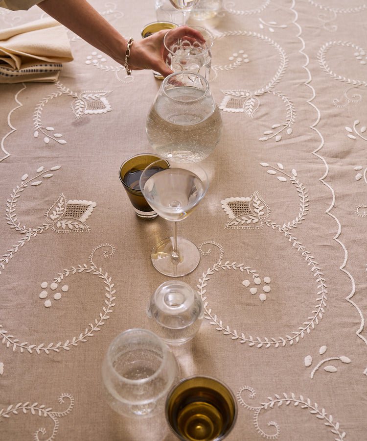 A hand arranges glassware and small cups filled with water on the Inez Tablecloth, crafted from raw linen and hand embroidered with traditional Portuguese designs, with a folded napkin in the corner.