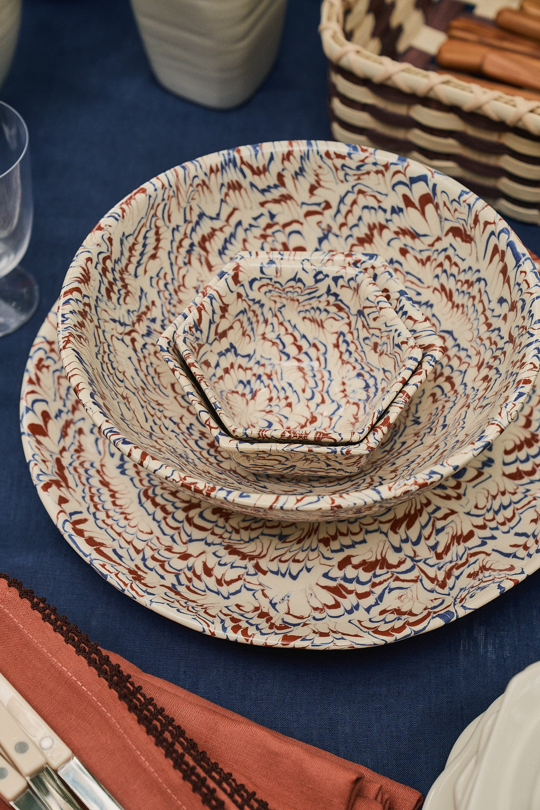 Three ceramic Celine Snack Bowls with marbled patterns are stacked on a blue tablecloth, next to glassware, cutlery, and a basket—evoking the timeless charm of classic Aptware, perfect for serving as elegant aperitivo bowls.