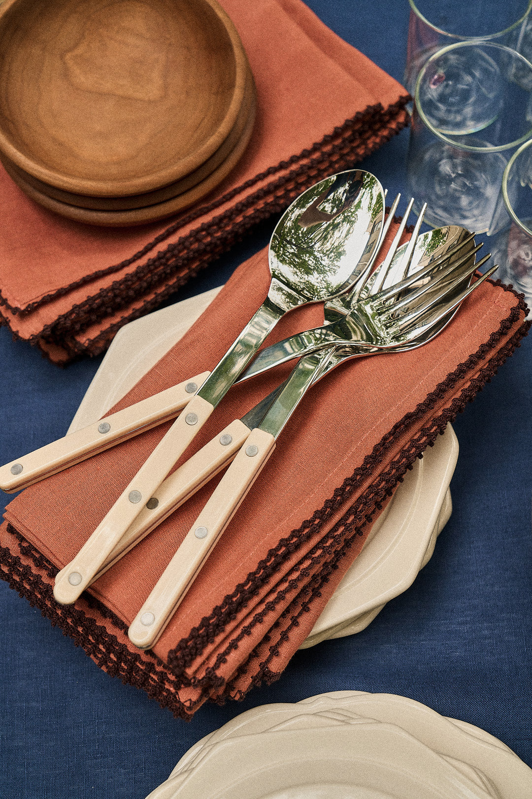 Three spoons and three forks with cream handles rest on Nico Napkins in rust, next to small plates, wooden bowls, and stacked glasses on a blue tablecloth.