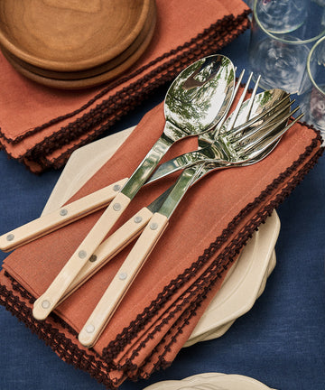 Three spoons and three forks with cream handles rest on Nico Napkins in rust, next to small plates, wooden bowls, and stacked glasses on a blue tablecloth.