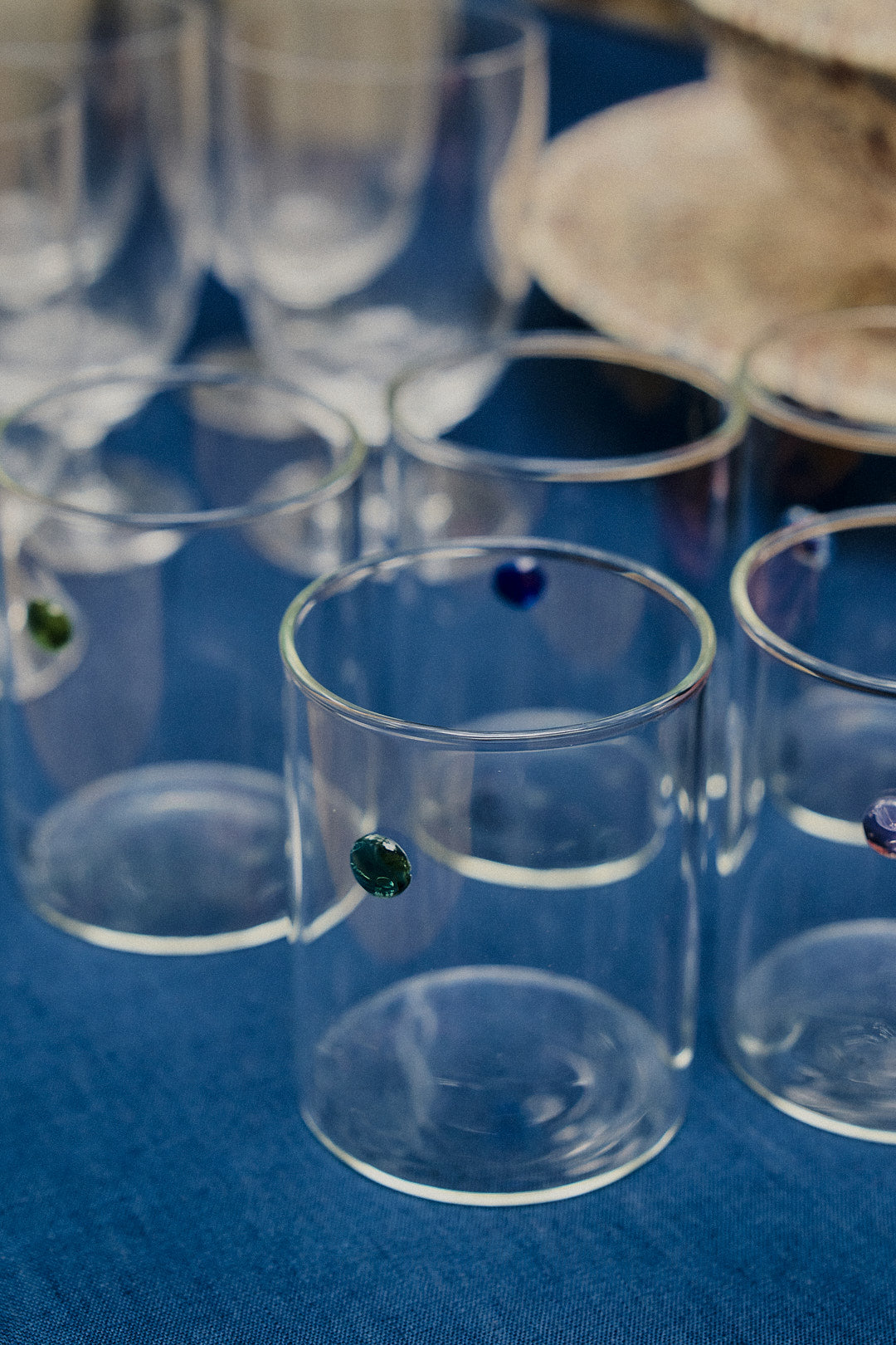 A set of Soleil Water Glasses, crafted from durable borosilicate glass using the lamp work technique and adorned with small colored dots, rests on a blue fabric surface. These contemporary tumblers mingle with other glassware and a beige item in the background.