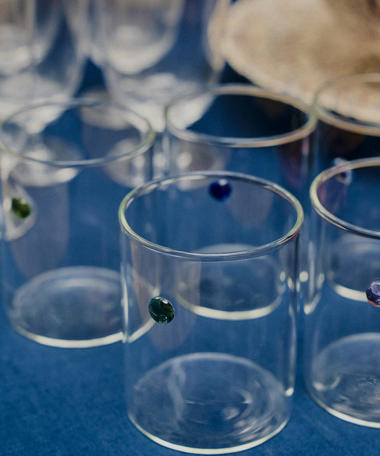 A set of Soleil Water Glasses, crafted from durable borosilicate glass using the lamp work technique and adorned with small colored dots, rests on a blue fabric surface. These contemporary tumblers mingle with other glassware and a beige item in the background.