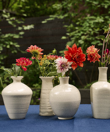 Six Cirrus Bud Vases, each crafted from white ceramic and designed to elegantly display vibrant flowers, are lined up on a blue tablecloth with green foliage in the background—a signature arrangement evocative of Frances Palmer’s artistry.
