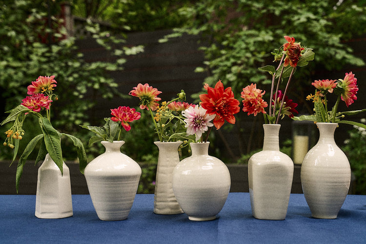 Six Cirrus Bud Vases, each crafted from white ceramic and designed to elegantly display vibrant flowers, are lined up on a blue tablecloth with green foliage in the background—a signature arrangement evocative of Frances Palmer’s artistry.