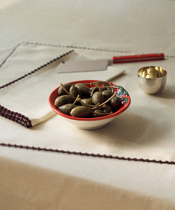 A Tomato Bowl sits on a table with a white cloth, next to a small metal cup, a folded napkin, and a butter knife—evoking the simple elegance of Italian farmhouse sideboard style.