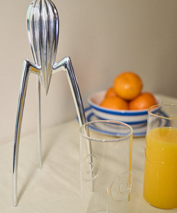 The Juicy Salif Citrus Squeezer, the iconic designer lemon juicer by Philippe Starck, stands as a striking modern kitchen accessory next to two clear glasses—one filled with fresh orange juice—with a bowl of oranges in the background on a light-colored surface.