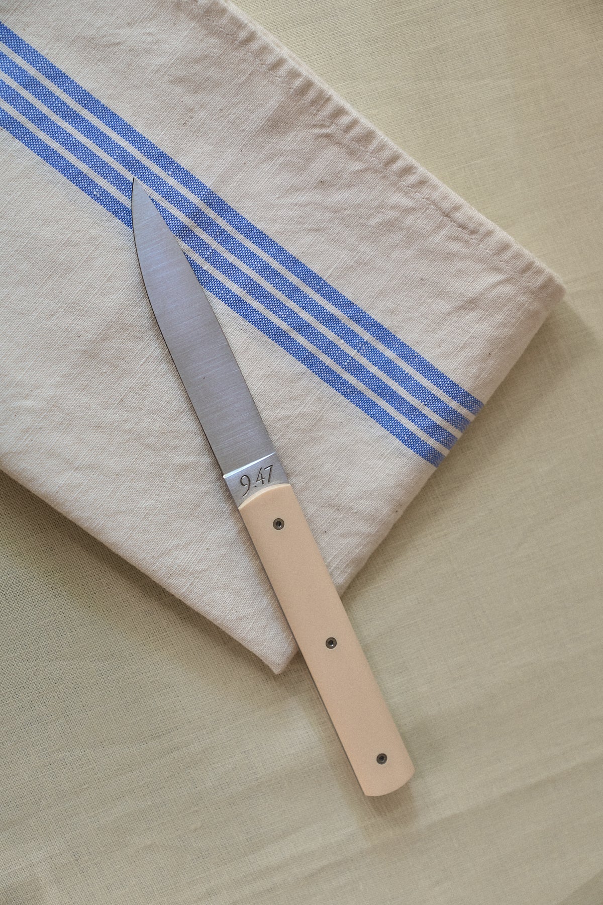 The Métis Tea Towel, with its beige fabric and blue stripes reminiscent of vintage designs, is folded on a light yellow surface, serving as the backdrop for a paring knife with a matching beige handle.