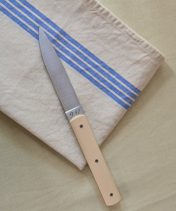 The Métis Tea Towel, with its beige fabric and blue stripes reminiscent of vintage designs, is folded on a light yellow surface, serving as the backdrop for a paring knife with a matching beige handle.