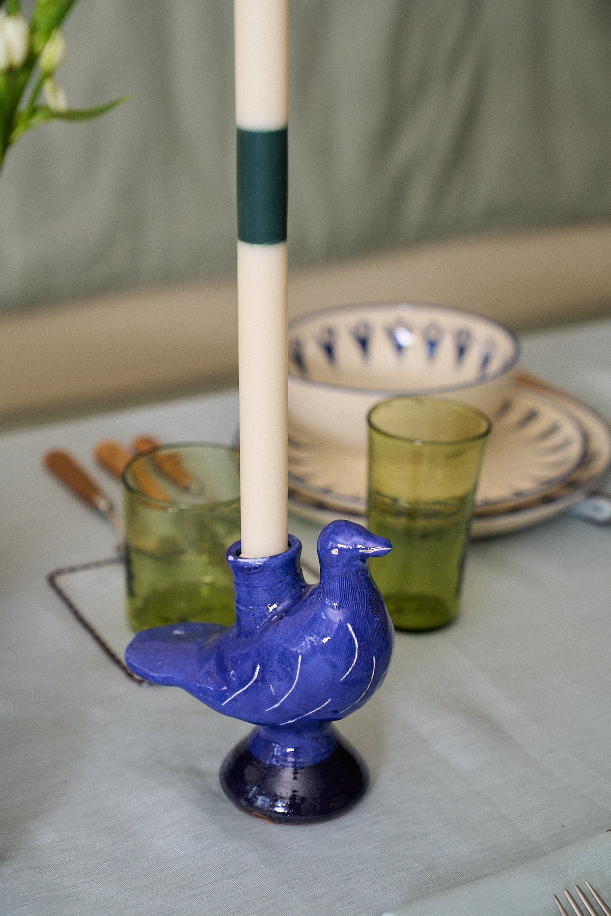 A white candle is placed in the Peace Dove Candle Holder, a blue ceramic piece with a vibrant cobalt glaze, on a set dining table alongside green glasses and patterned plates.