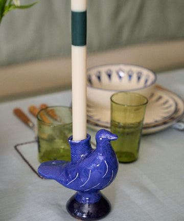 A white candle is placed in the Peace Dove Candle Holder, a blue ceramic piece with a vibrant cobalt glaze, on a set dining table alongside green glasses and patterned plates.