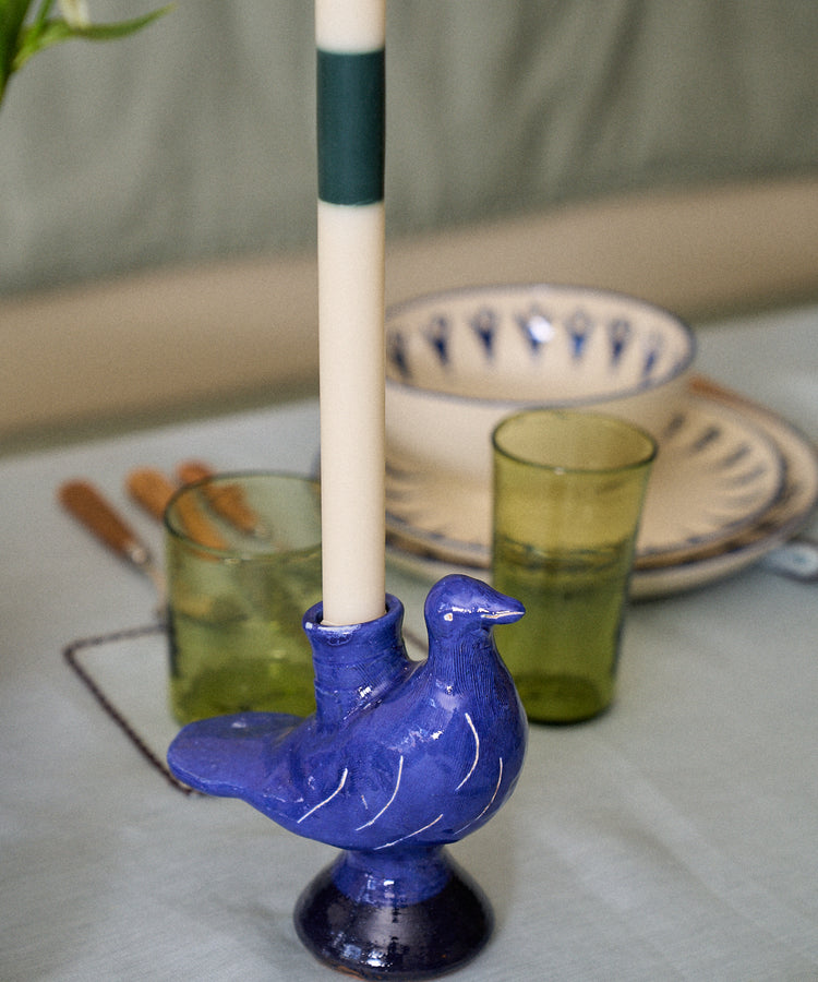 A white candle is placed in the Peace Dove Candle Holder, a blue ceramic piece with a vibrant cobalt glaze, on a set dining table alongside green glasses and patterned plates.