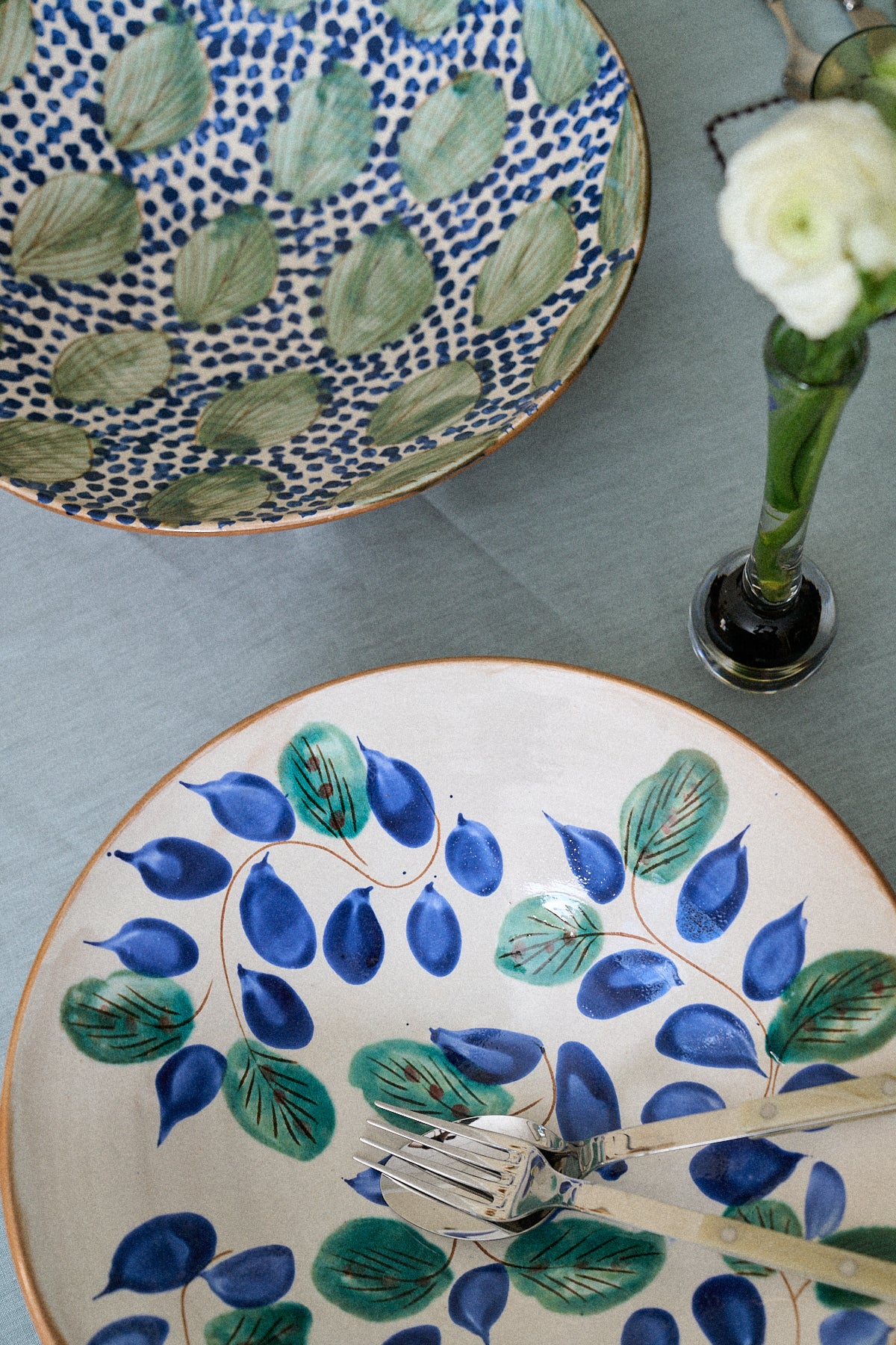 A Midnight Garden Serving Bowl rests on a light blue tablecloth next to two forks and a single white flower in a glass vase.