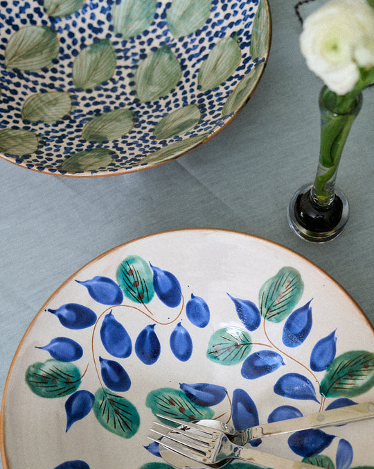 A Midnight Garden Serving Bowl rests on a light blue tablecloth next to two forks and a single white flower in a glass vase.