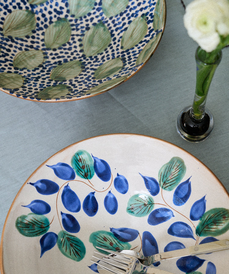 A Midnight Garden Serving Bowl rests on a light blue tablecloth next to two forks and a single white flower in a glass vase.