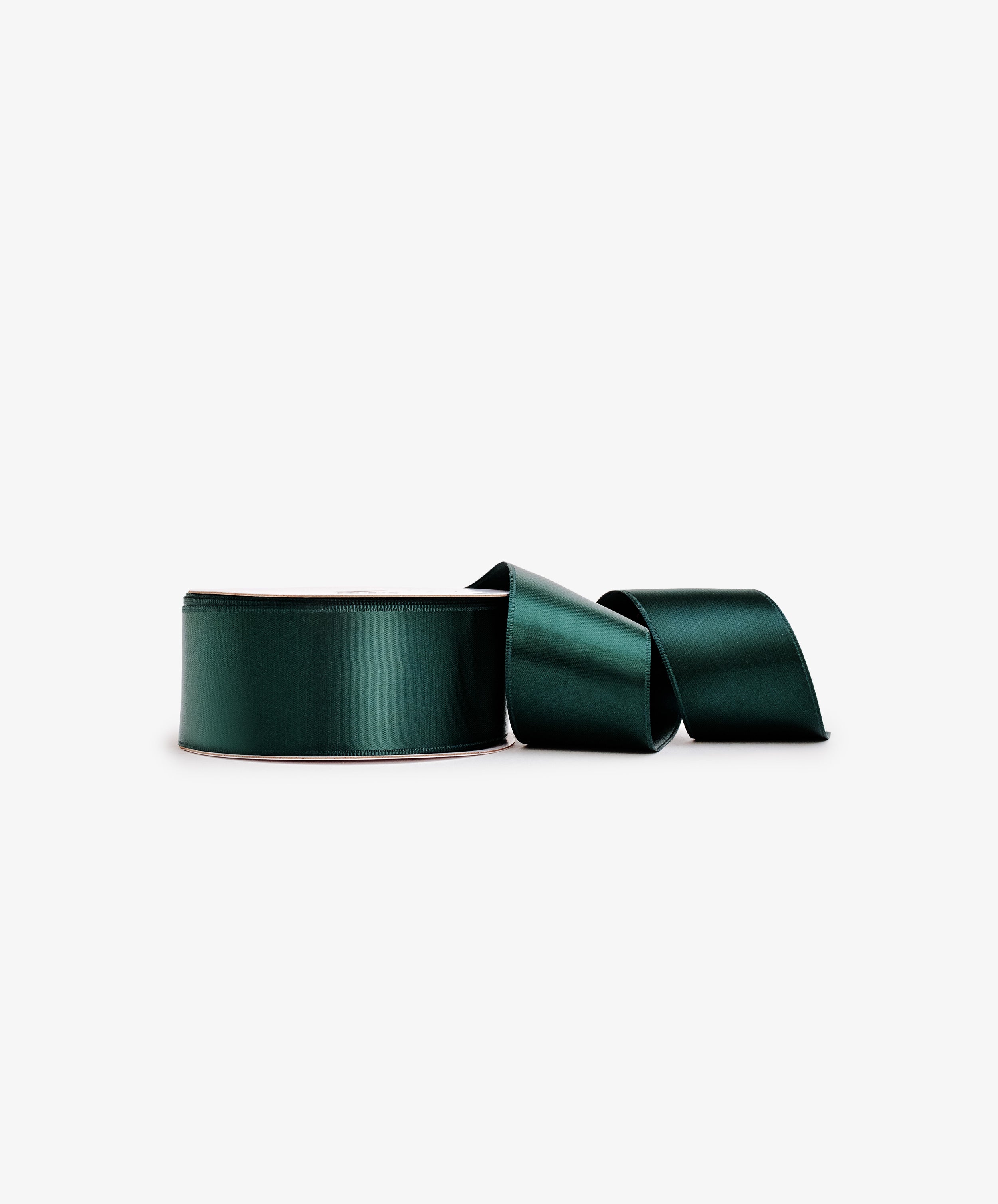 A partially unspooled roll of Double Faced Satin Ribbon in dark green rests on a plain white background, ideal for elegant gift wrapping.