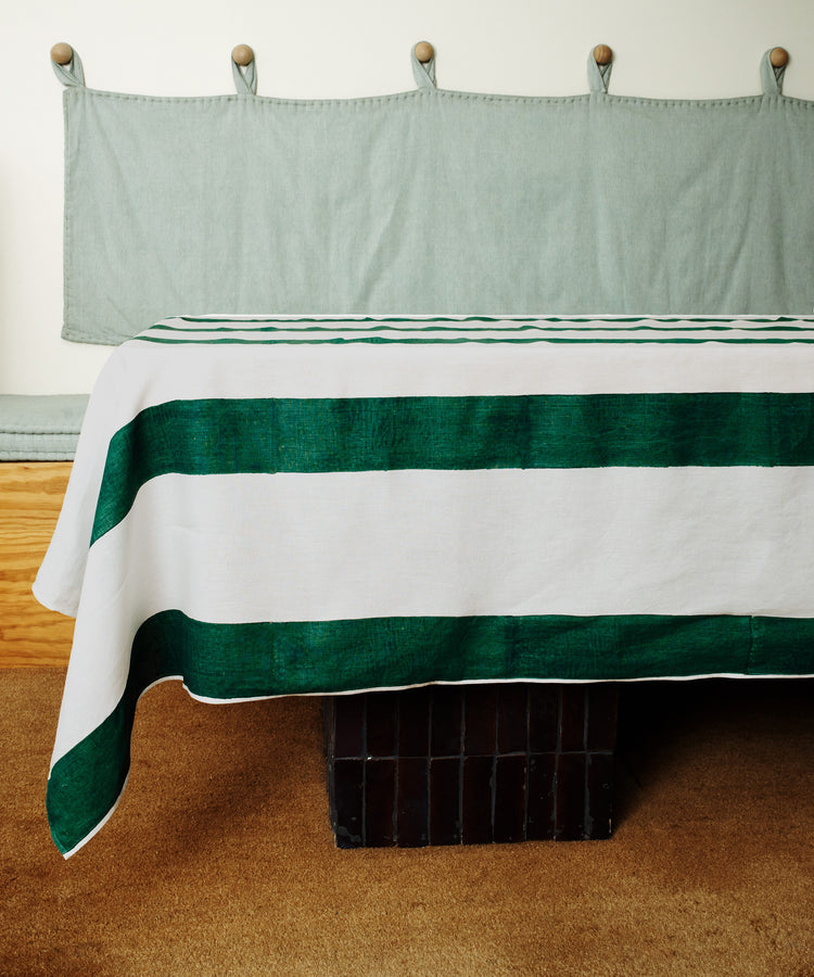 A table with a hand-printed green-striped Riga Tablecloth stands on a brown carpet, with a light green bench and cushion along the wall in the background.