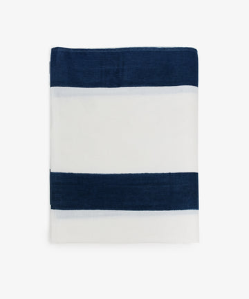 The Riga Tablecloth, hand-printed with two horizontal navy blue stripes on indigo-striped linen, is folded neatly against a plain white background for an artisanal touch.
