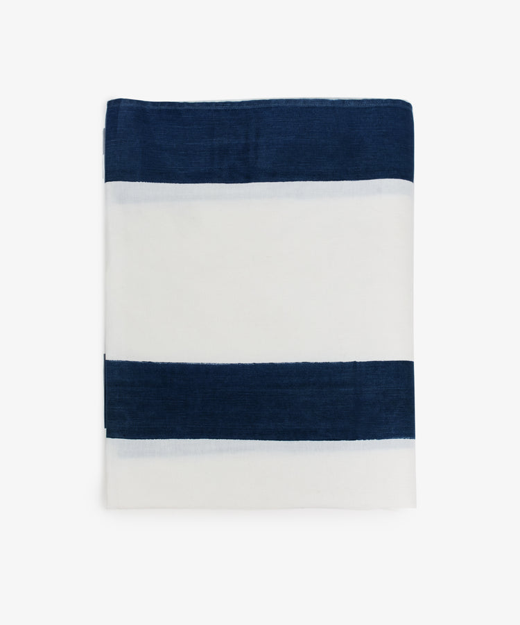 The Riga Tablecloth, hand-printed with two horizontal navy blue stripes on indigo-striped linen, is folded neatly against a plain white background for an artisanal touch.